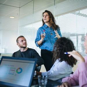 Businesswoman walking in office boardroom and sharing a strategy with team sitting at table. Female manager explaining new business plan with coworkers sitting at conference table in office.